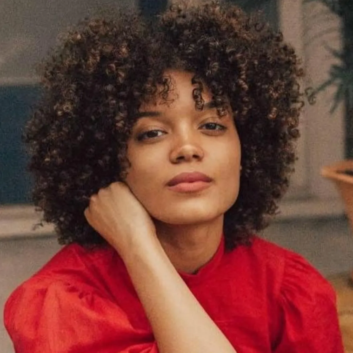 A woman with curly hair wearing a red top poses thoughtfully, resting a hand on their chin, in a warmly lit room with blurred background elements.