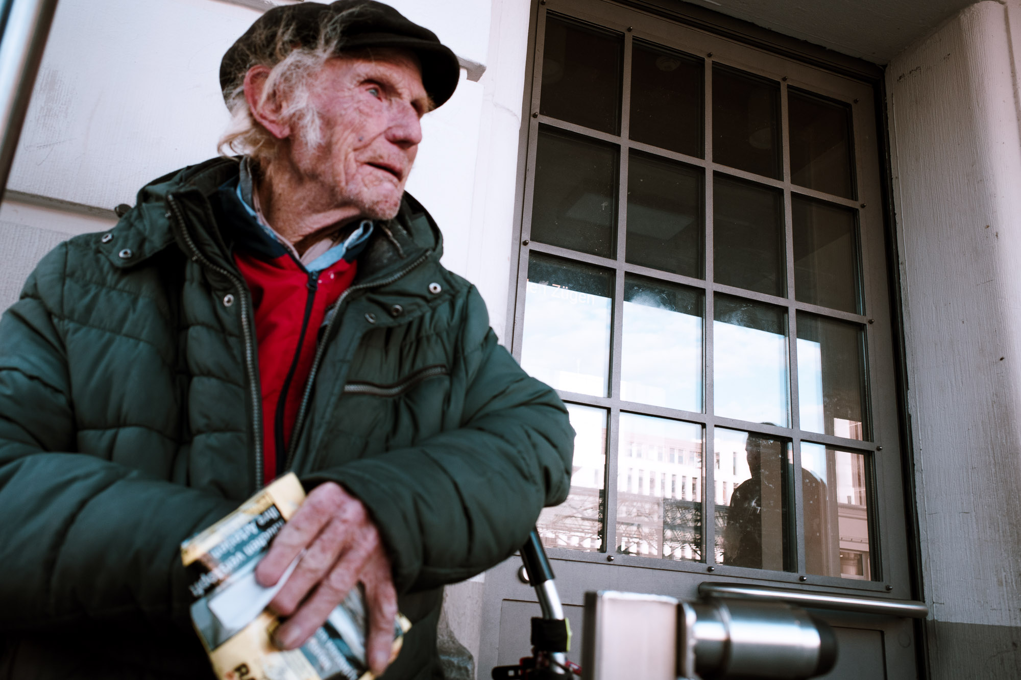 An elderly man wearing a green puffer jacket and black cap stands outside, clutching a book, with a reflective window and urban backdrop.