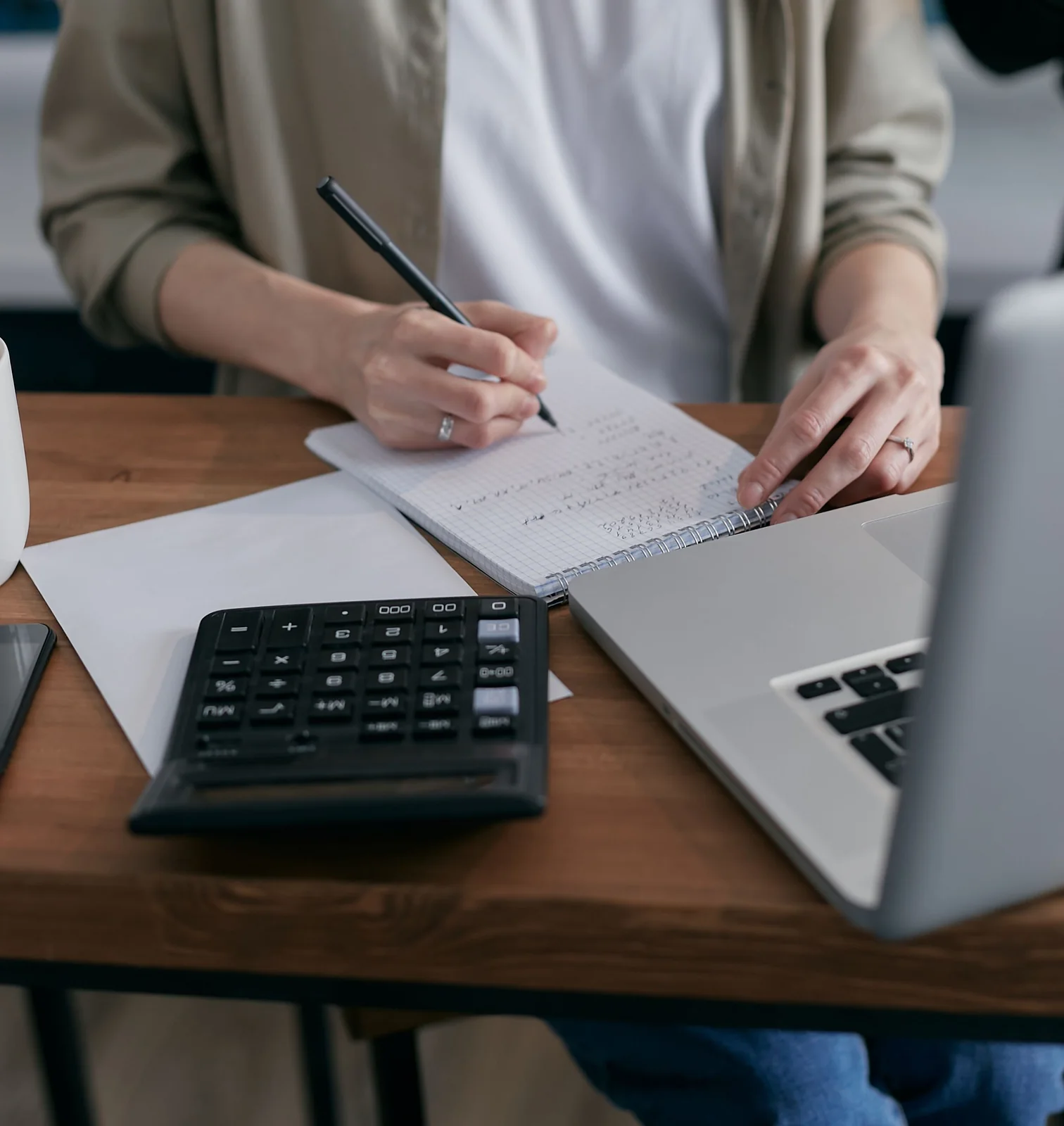 A person writes budget calculations in a notebook beside a calculator.