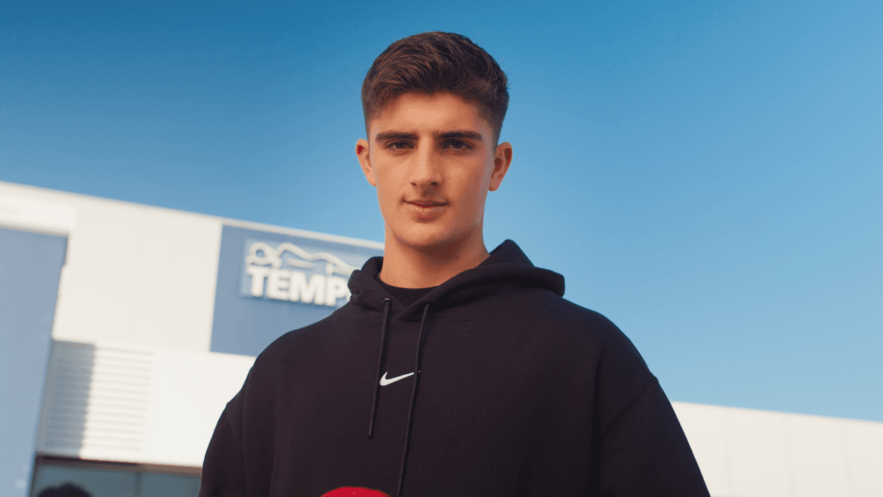 A young man with short dark hair wearing a dark hoodie stands outdoors, with a blue sky visible in the background