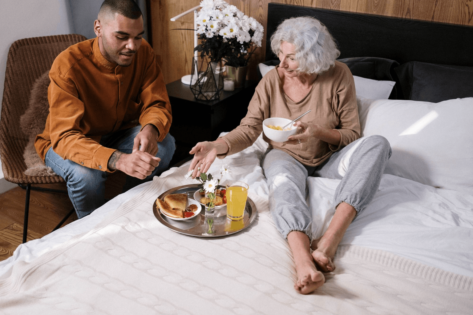 A young man bringing breakfast on a tray to the older woman he looks after at home.