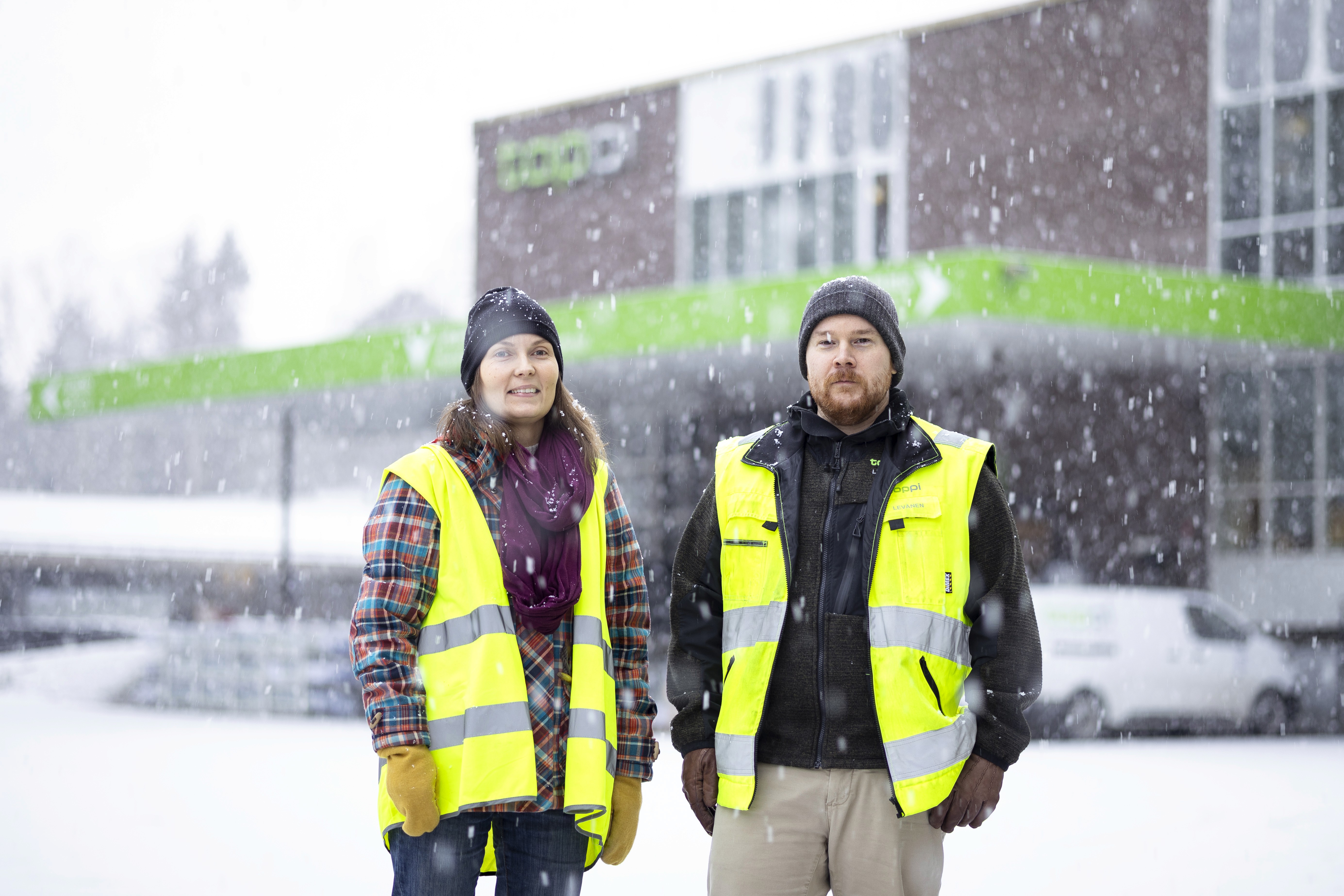 Two individuals in yellow safety vests stand in a snowy landscape, with a building in the background