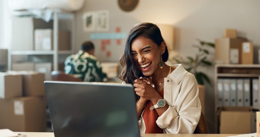 Smiling woman working on a laptop at a kitchen table with a cup of coffee.