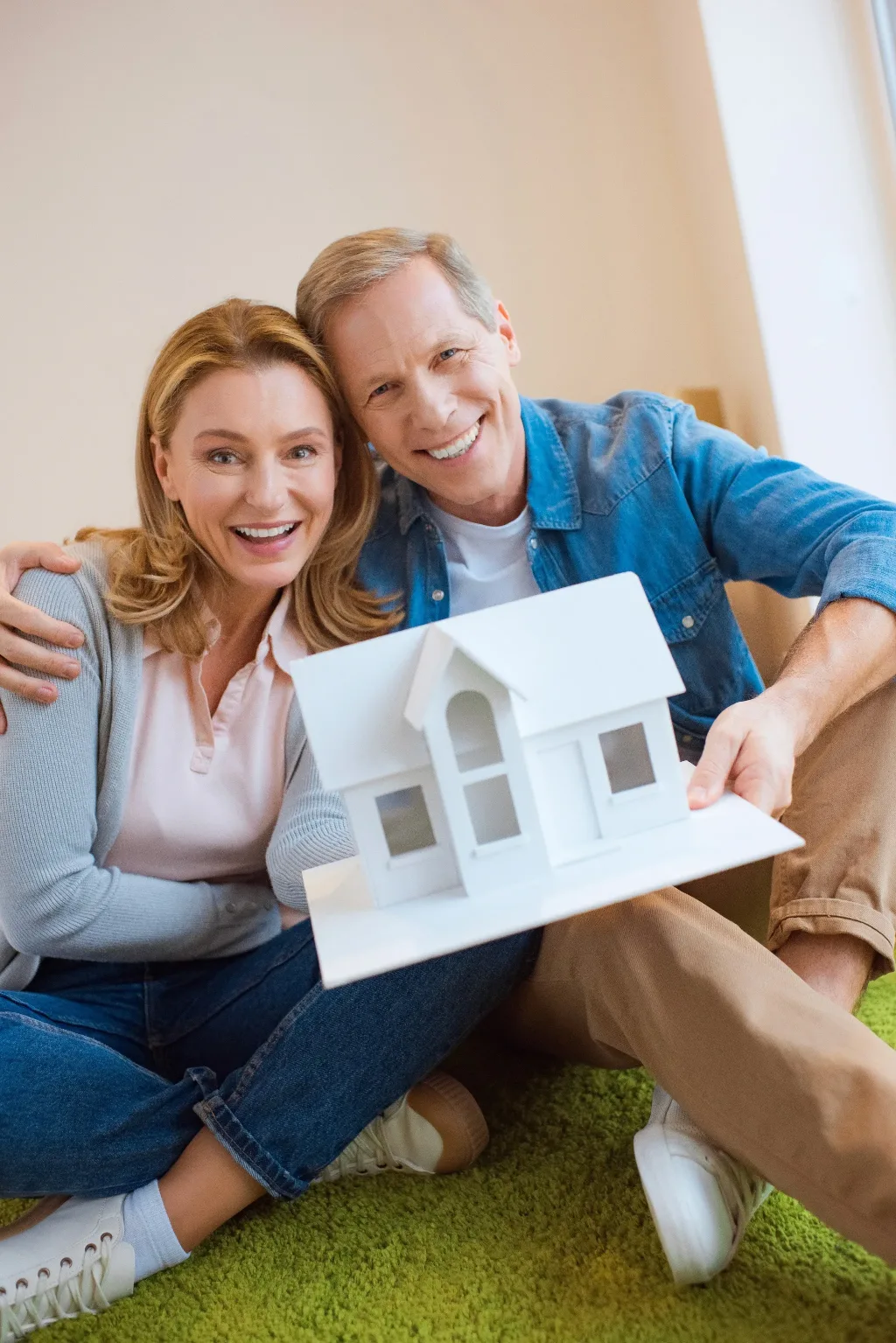 A smiling middle-aged couple sits together on a green carpet holding a white miniature house model.