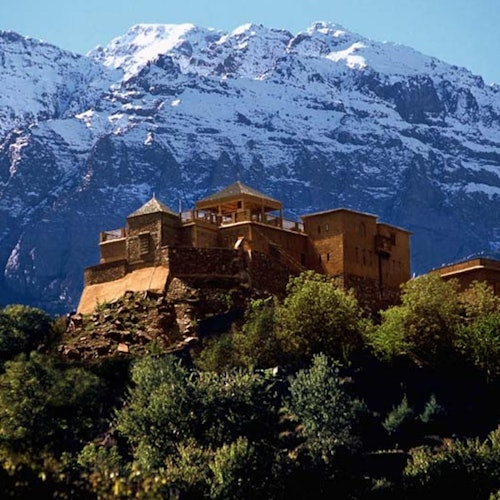 A stone building with multiple levels sits atop a hill with green foliage, against a backdrop of snow-capped mountains.