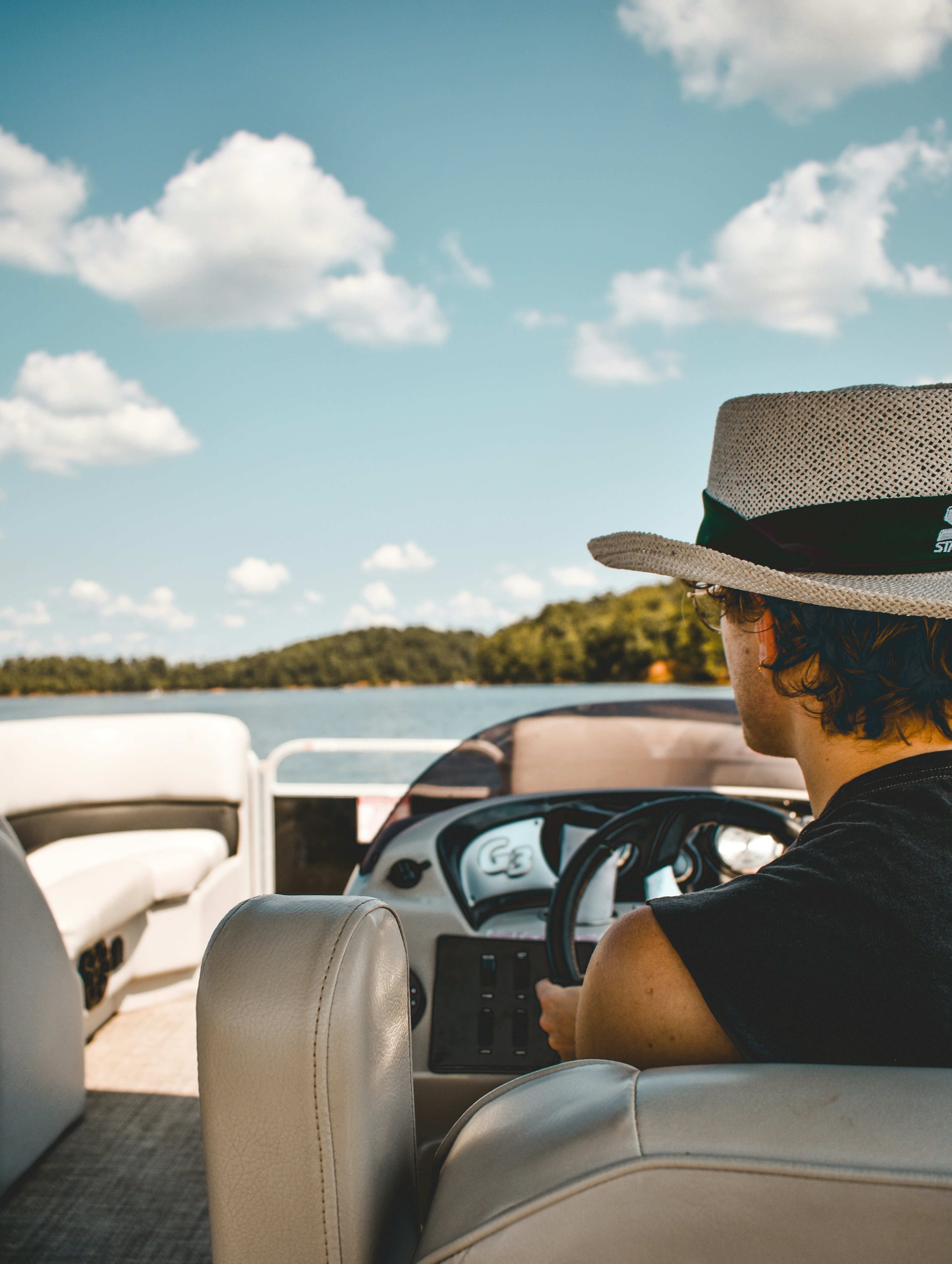 A person wearing a hat is steering a boat on a tranquil lake under a partly cloudy sky, surrounded by lush green trees in the background.
