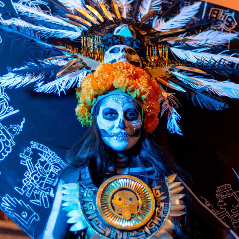 Fotografía de una mujer en su rol de madre y danzante, portando indumentaria tradicional mexica que incluye un penacho de plumas y pintura facial ritual, capturando el momento previo al inicio de una ceremonia de danza.