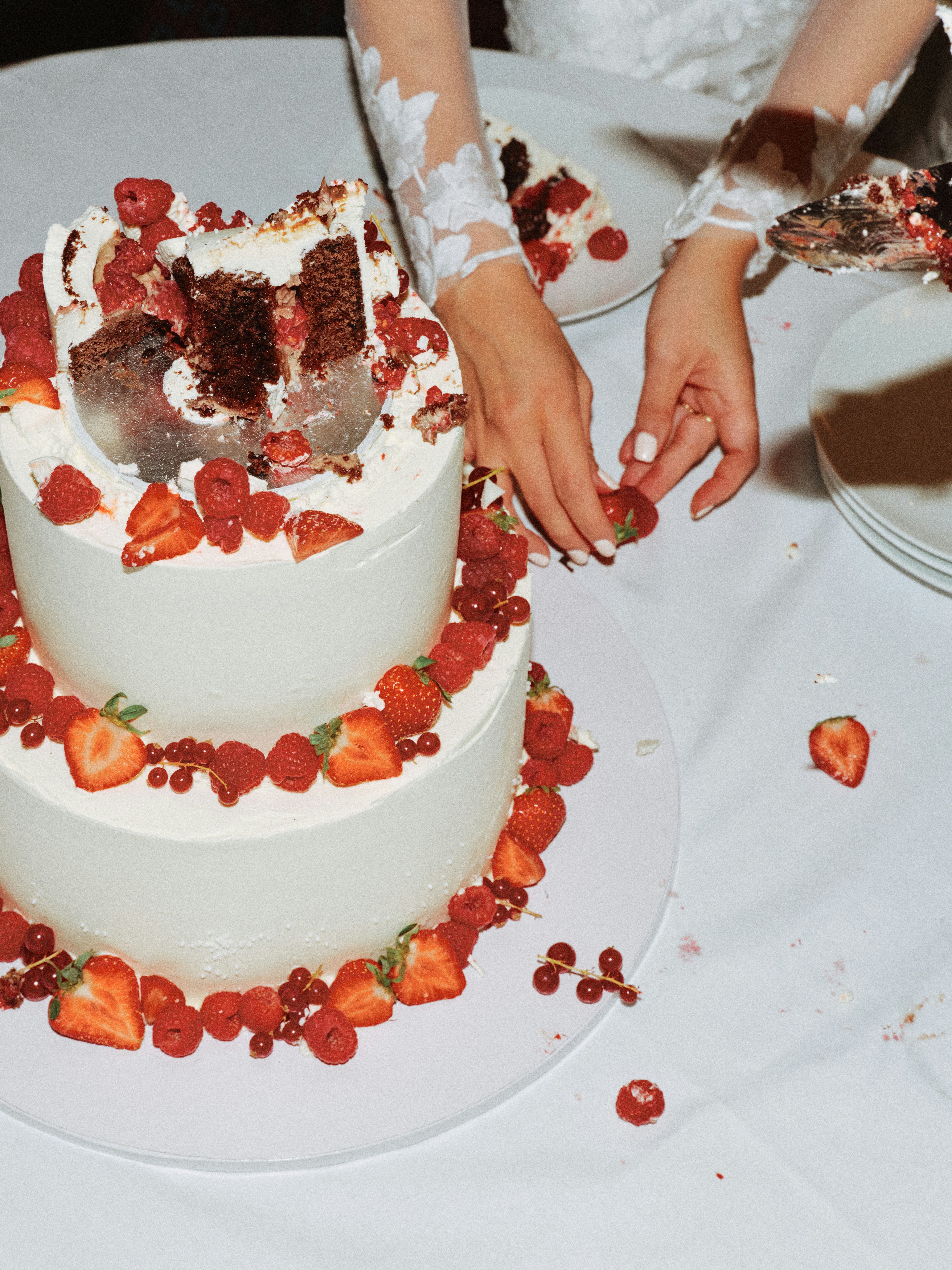 Close-up of a bride's hands in a lace embroidered wedding dress picking strawberries from a partially cut three-tier white wedding cake decorated with fresh red berries