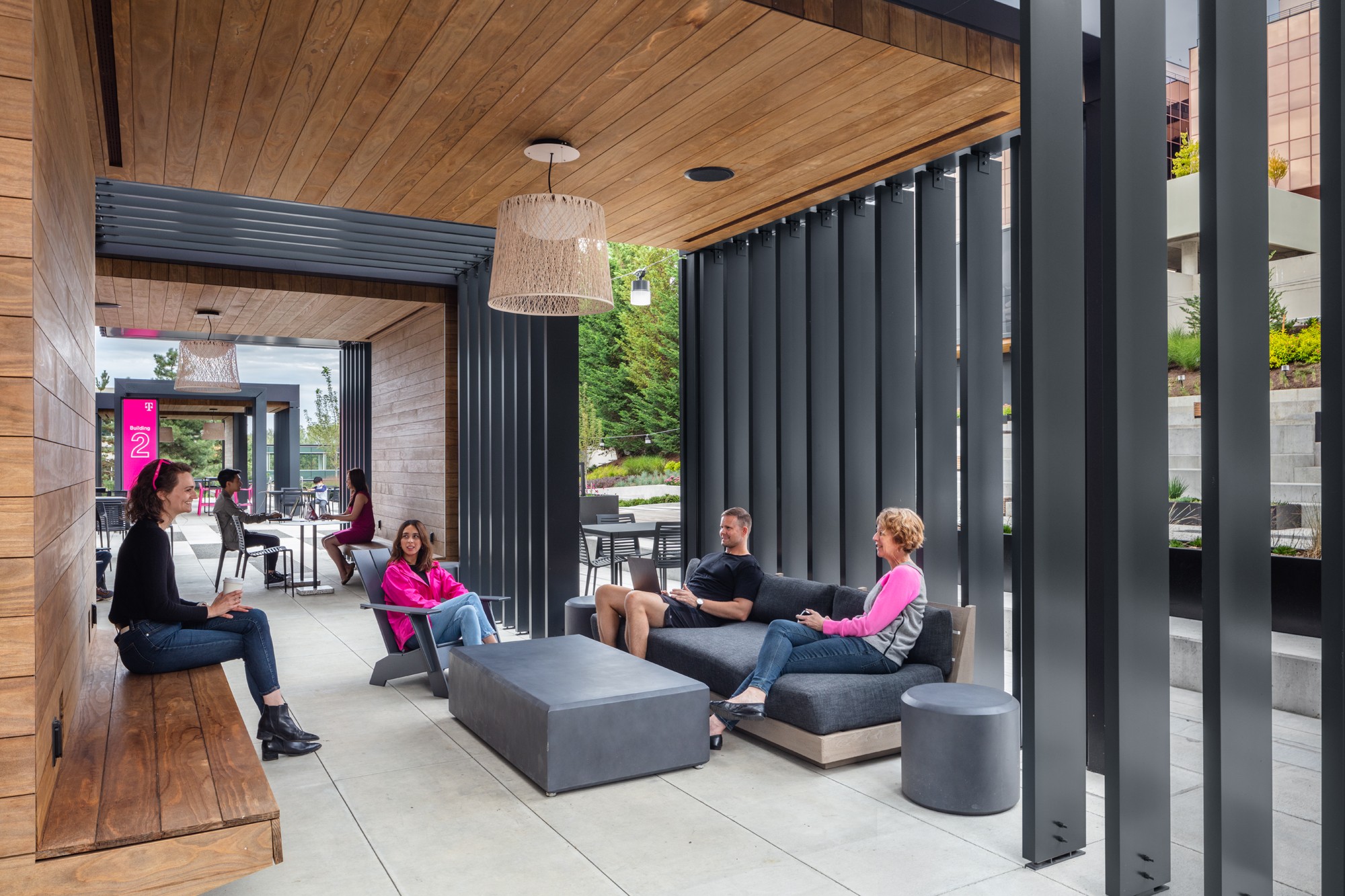 Individuals relax on sofas in the completed digit pavilion, a modern outdoor lounge with a warm wooden wall and ceiling paired with sleek metal fins, perfectly reflecting the concept renderings.