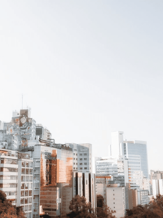 A couple is lying on a bed surrounded by moving boxes and suitcases, looking lovingly into each other's eyes. The man has long dark hair and a beard, and the woman has curly blonde hair, wearing a pink shirt tied at the waist.