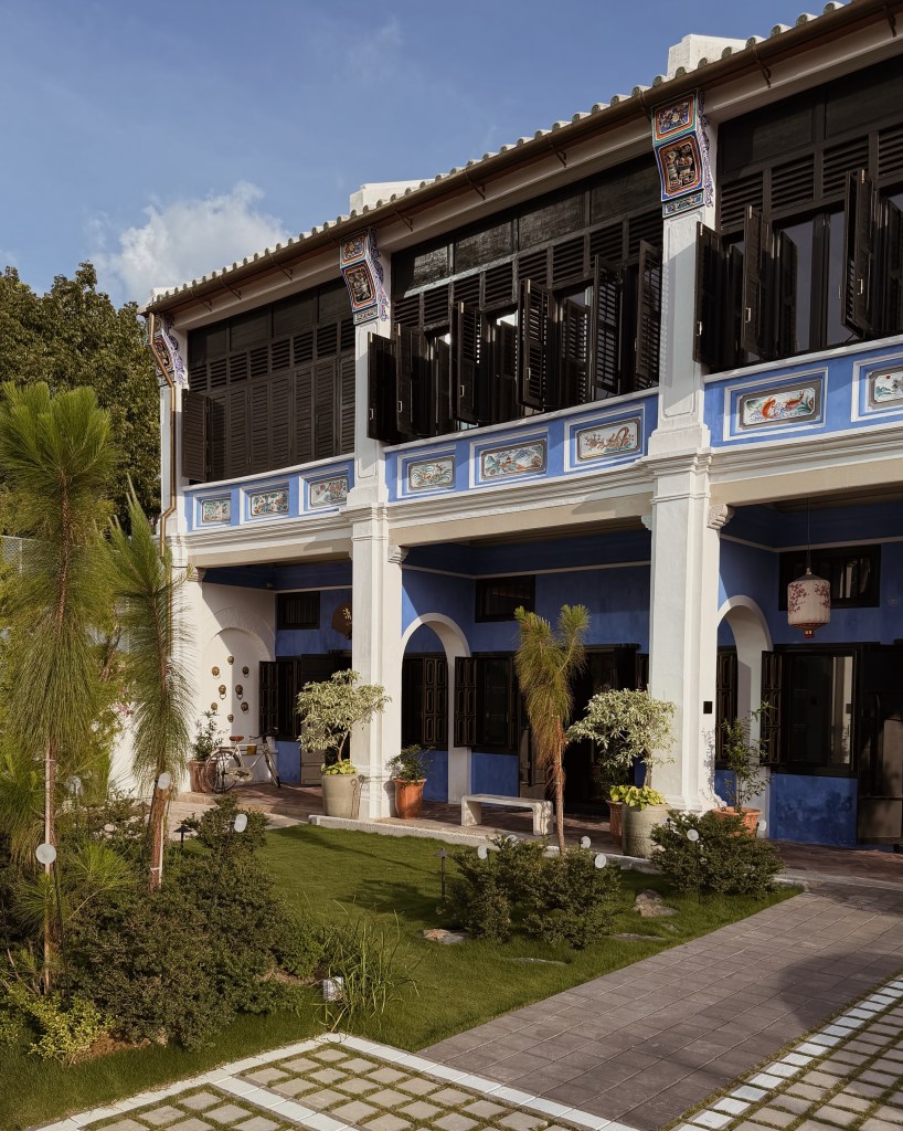 A charming colonial-style building with blue walls, decorative panels, and open shutters, surrounded by a lush garden featuring various plants and a stone pathway.