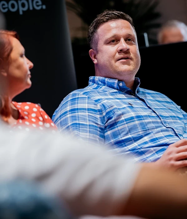 Two people seated side by side during an indoor discussion or panel event. One person is wearing a blue checkered shirt and is slightly turned toward the other, holding their hands together in their lap. The other person is wearing a red top with a white polka‑dot pattern. A dark backdrop and event signage are visible behind them, suggesting a professional talk or community discussion setting.