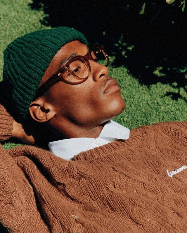 Profile of a person wearing a green knit beanie and brown glasses, lying in the grass with eyes closed in bright sunlight.