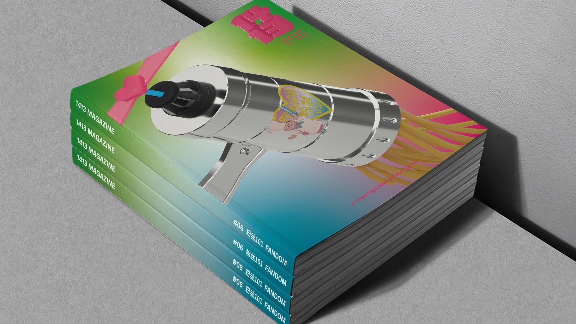 A stack of magazines rests on a gray surface. The top magazine displays a silver cylindrical device with a black nozzle and a pink ribbon tied around it. The cover transitions from green to teal, with a pink and yellow gradient. The bottom magazine shows the same design but with green and teal tones. The spine of each magazine displays “1413 MAGAZINE” vertically.