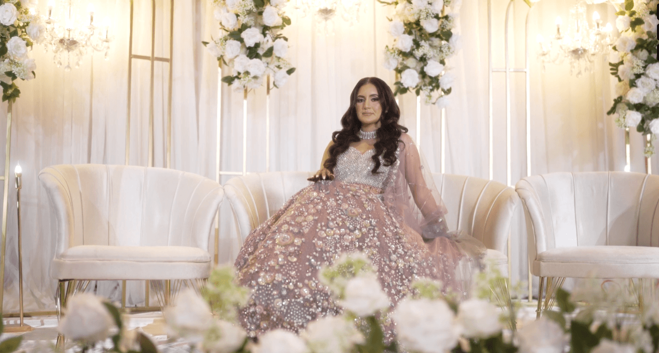 Woman in a pink dress sitting on a white chair, surrounded by elegant lighting, white and off-white decor, and flowers, looking at the camera in a beautifully styled room.