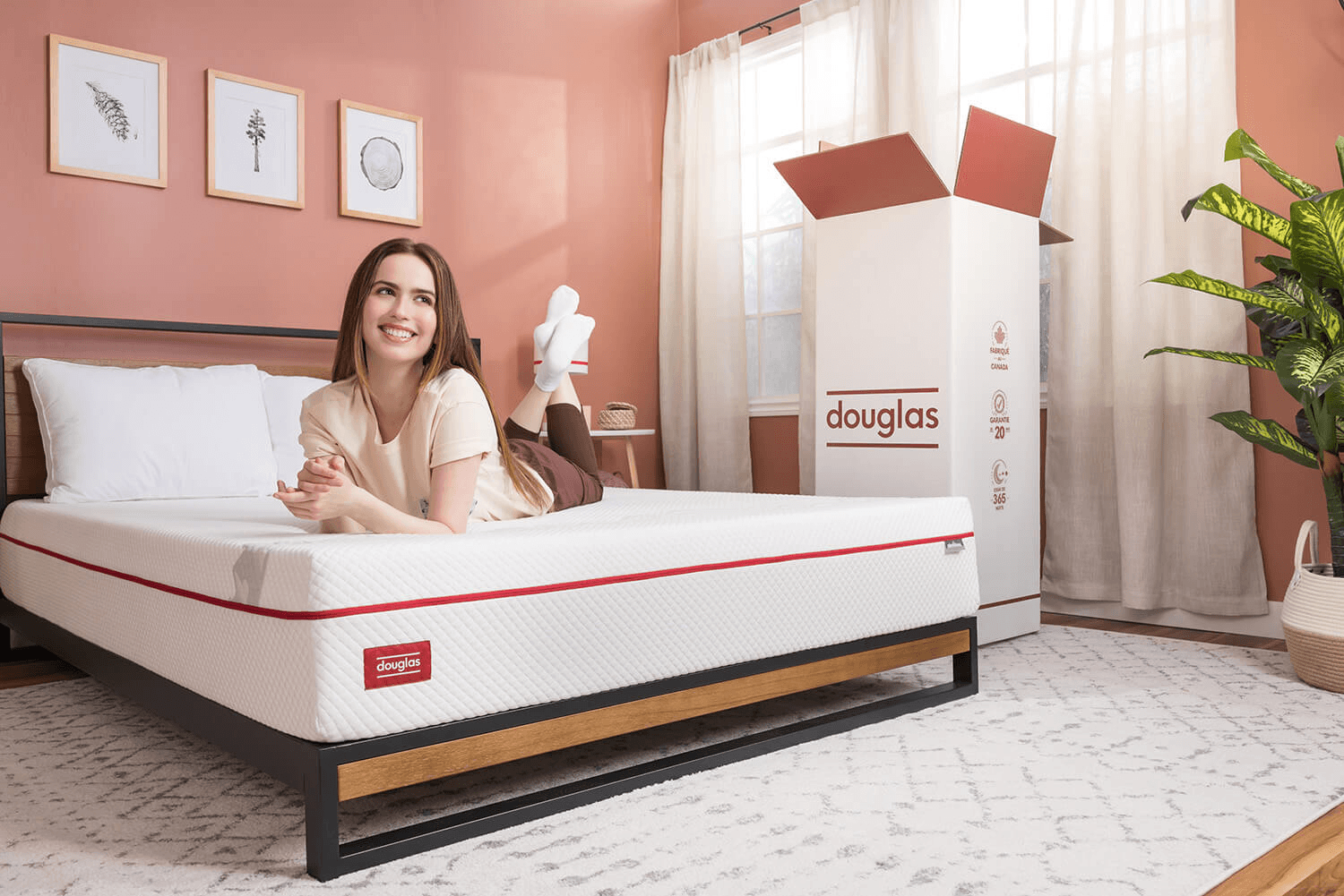 A woman smiling while lying happily on a Douglas Original mattress on a bed frame. The mattress features its red border trim and Douglas label. In the background, a large cardboard Douglas mattress-in-a-box stands upright, illustrating doorstep delivery to a Canadian apartment.