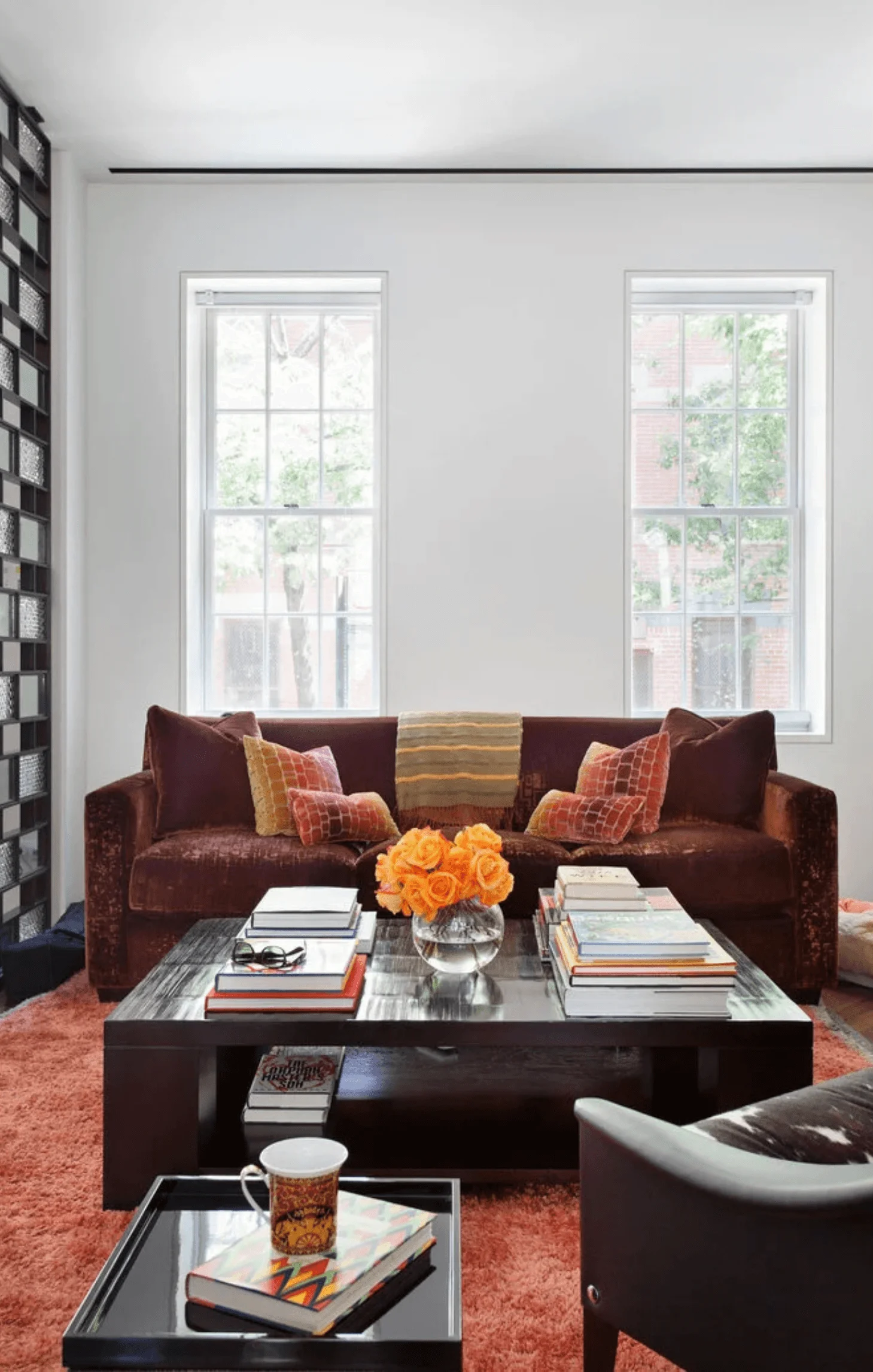 A deep brown velvet sectional sofa sits on a striped rug, with colorful throw pillows and stacks of books on the coffee table