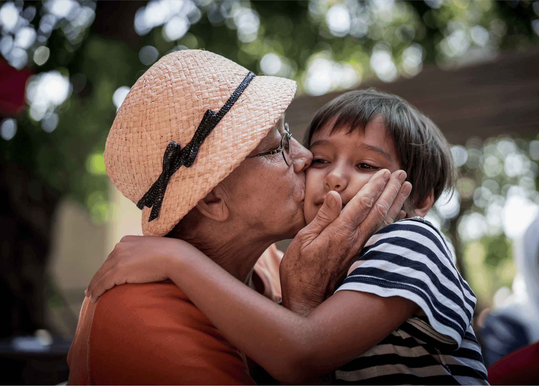 An elderly person in a straw hat lovingly kisses a young child wearing a striped shirt, set against a blurred outdoor background with green foliage and dappled sunlight.