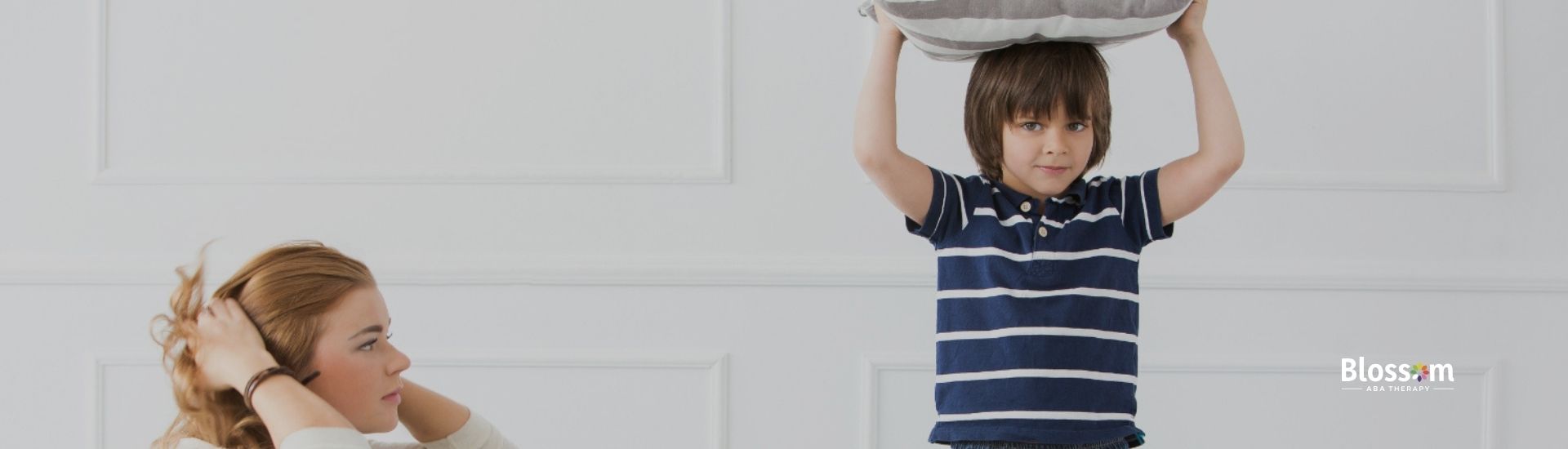 A boy standing on a couch, holding a pillow above his head while his mother looks frustrated.