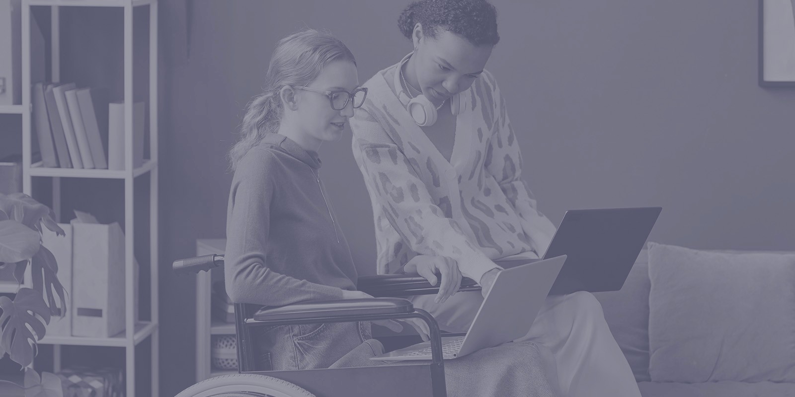 A young woman in a wheelchair and another woman sitting on a sofa while collaborating together on their laptops