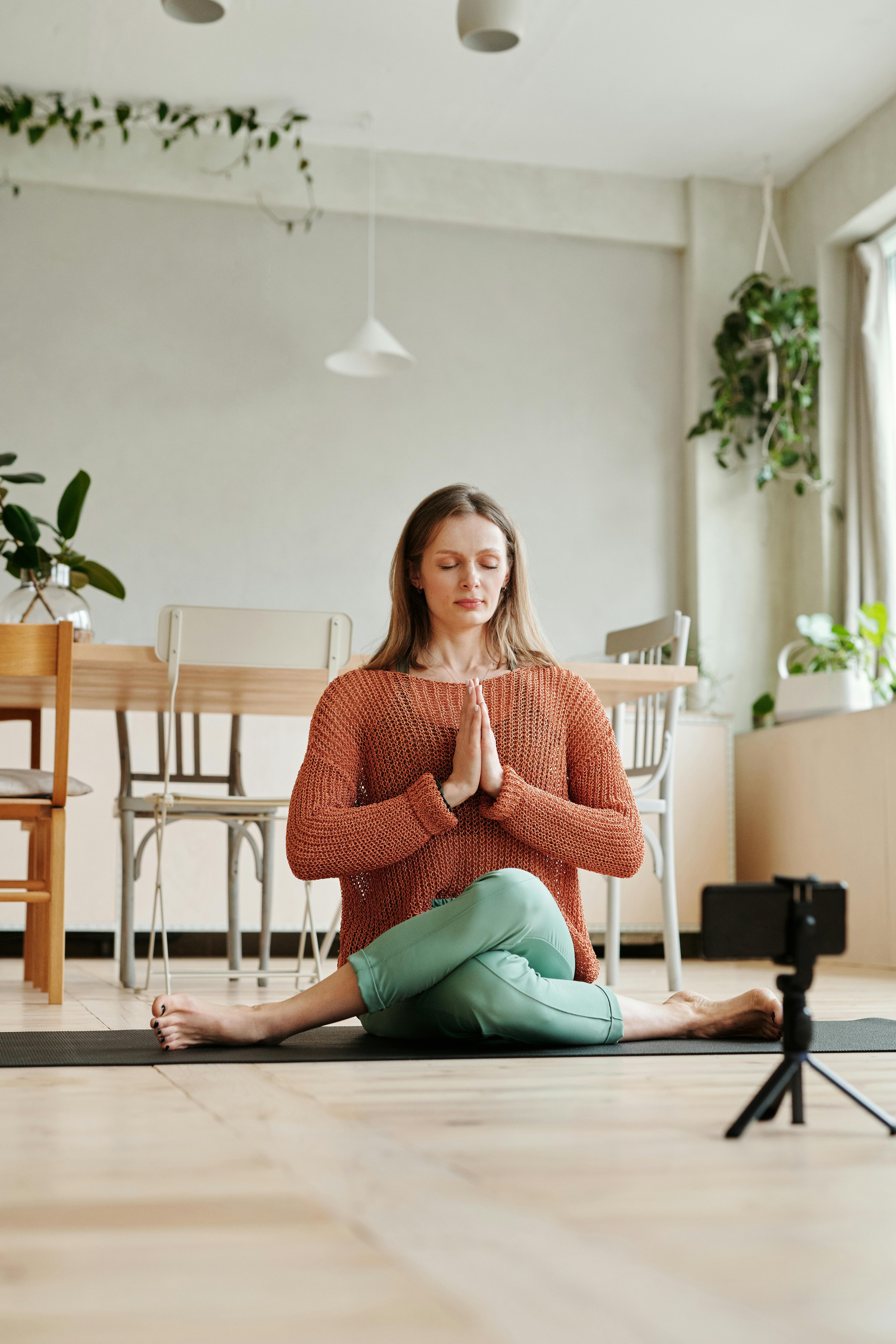 A person practices yoga indoors, seated cross-legged with eyes closed and hands in prayer position. The room is calm and light-filled, with plants and furniture in the background. A small tripod is set up nearby.
