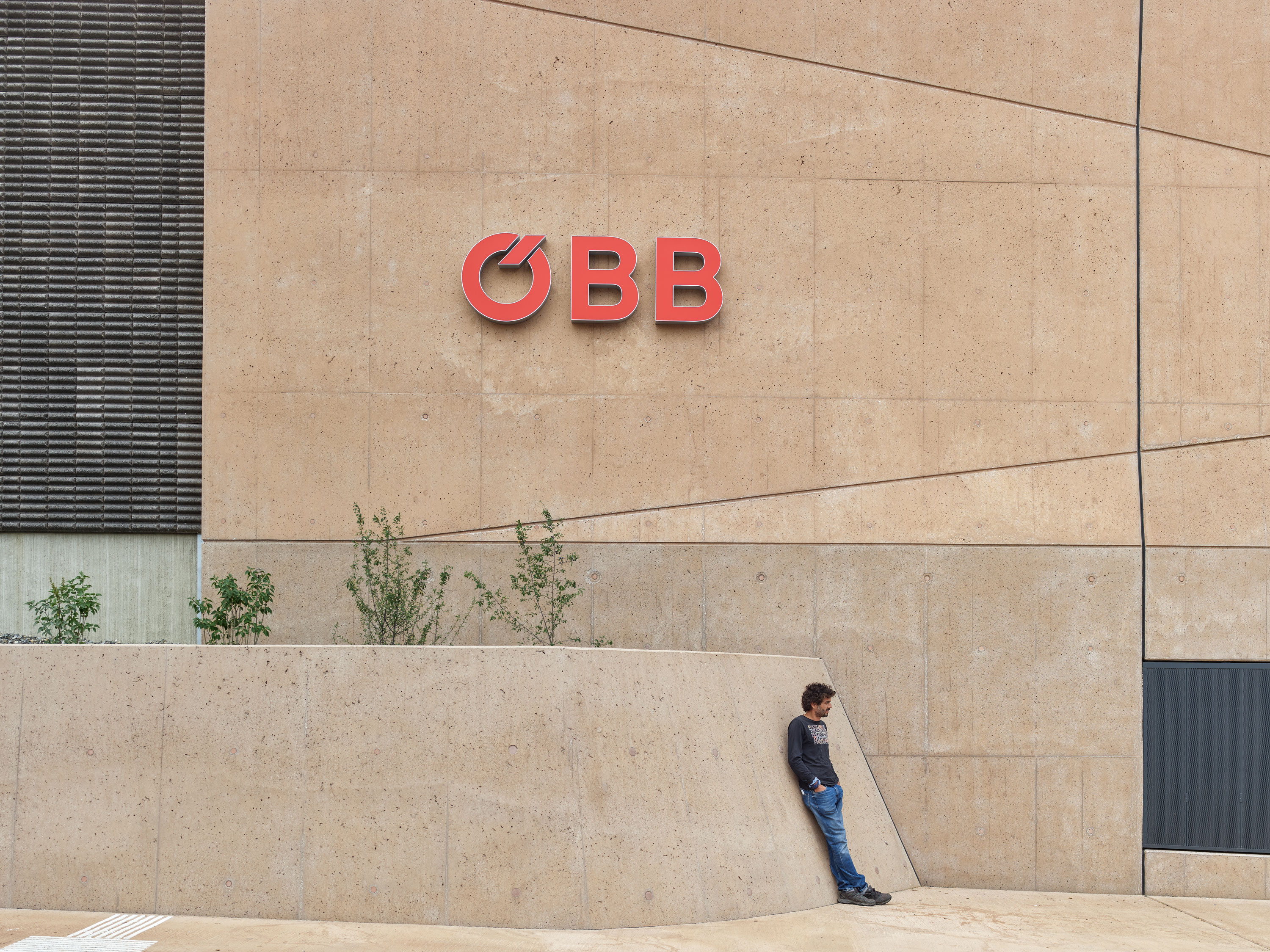 Mann lehnt an einer hellen Betonwand mit rotem ÖBB-Logo, umgeben von Pflanzen.