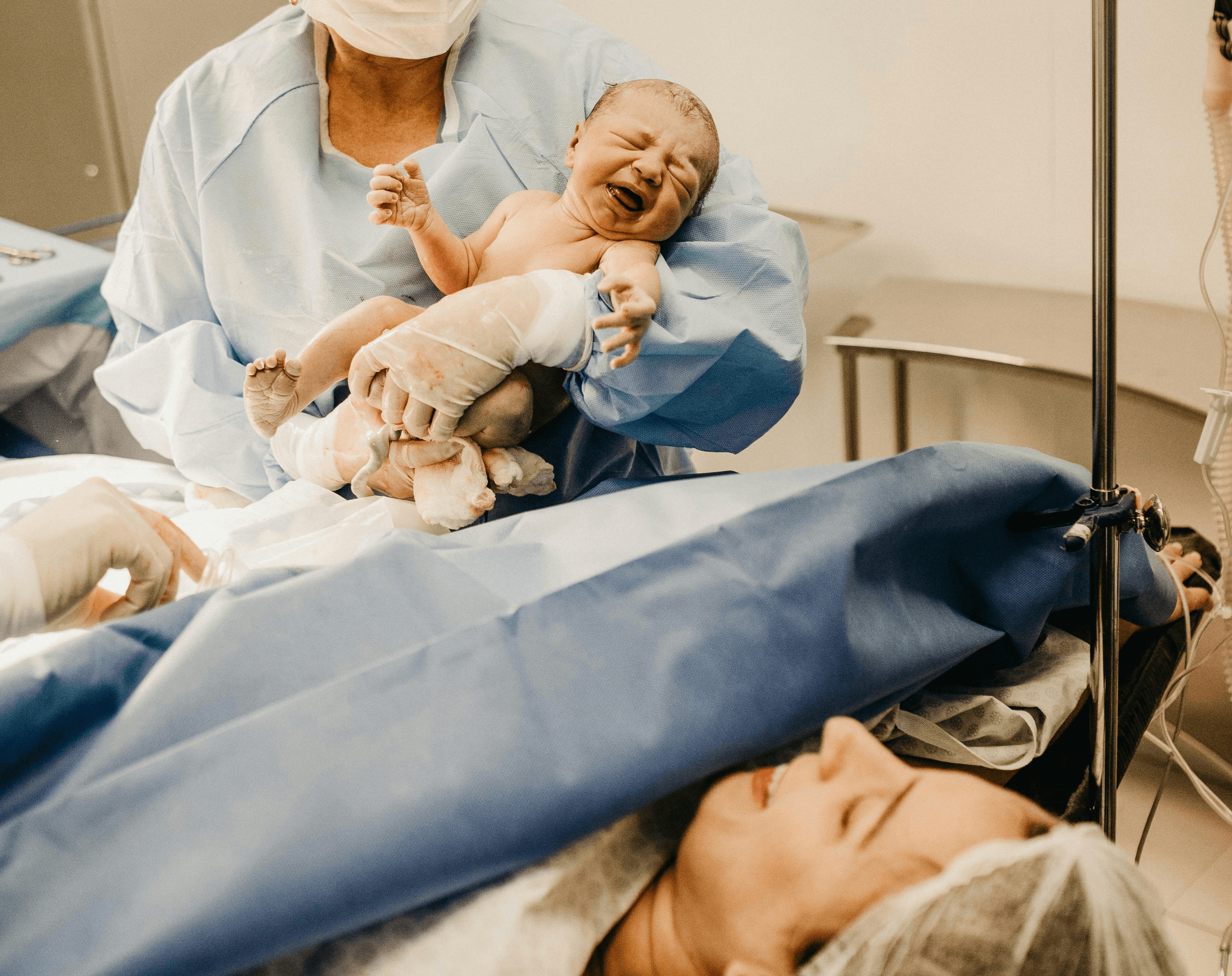 A medical practioner holds a newborn baby delivered through caesarean section and is lifting it towards the mother. In focus there is the mother's head, she is lying on the operating table with a sheet up in front of her face.