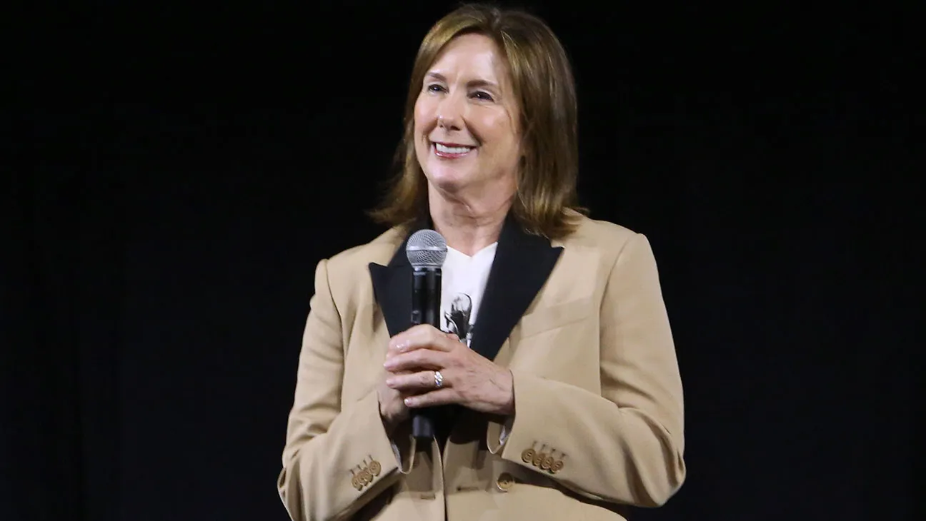 Lucasfilm President Kathleen Kennedy holding a microphone wearing a brown jacket in front of a black background