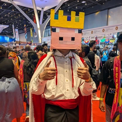 A photograph of a person at a crowded indoor convention cosplaying as the Minecraft character Technoblade. They are wearing a large, blocky, pink cardboard mask resembling a Minecraft pig, topped with a yellow blocky crown featuring blue and green square jewels. They are dressed in a white long-sleeved button-up shirt, black pants, a wide red fabric sash around their waist, and a long red cape with white fuzzy trim fastened at the neck with a gold chain. The cosplayer is standing on a red convention carpet and giving a thumbs-up gesture with both hands. The background is a busy exhibition hall with a high ceiling, large white structural pillars, various publisher booths, and a large crowd of attendees walking around.