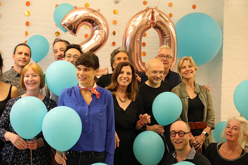 A photo of a group of people surrounded by blue balloons and in the background the number twenty made also from shiny balloons.