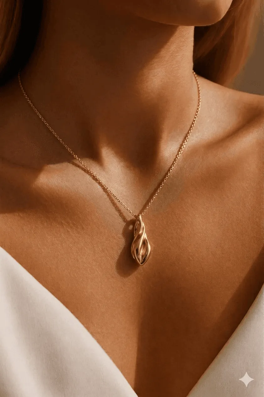 A woman wearing a minimalist white outfit showcases a delicate gold necklace with an elegant teardrop pendant, set against a softly lit background.