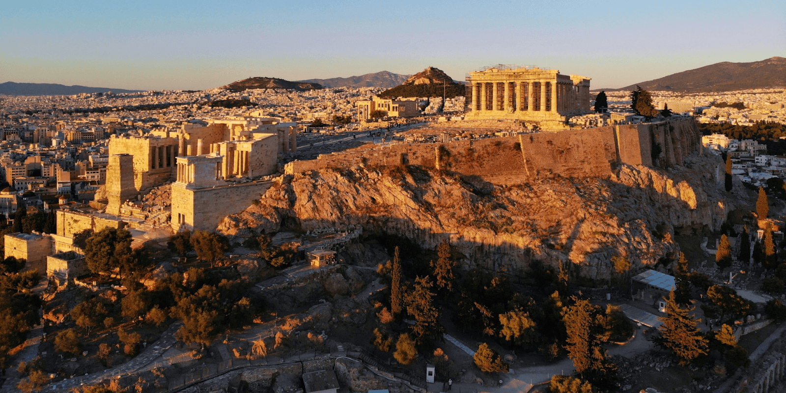 View of the Acropolis in Athens at sunset, with the Parthenon illuminated above the city and surrounding hills.