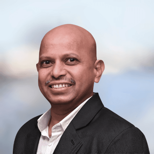 A smiling man with curly hair is sitting at a desk, looking at the camera, with a laptop and a lamp in the background.