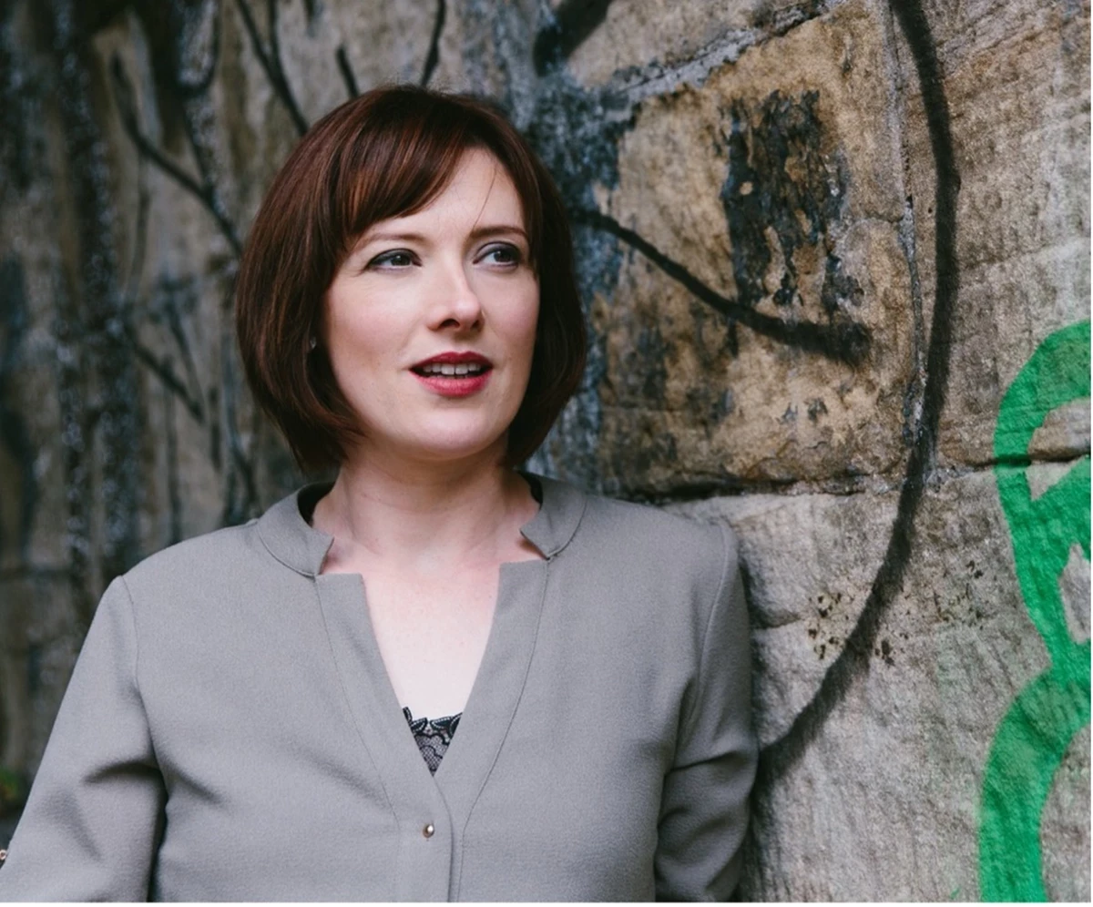 A person wearing a light grey blouse stands outdoors beside a stone wall covered with graffiti. The setting has a textured, urban feel.