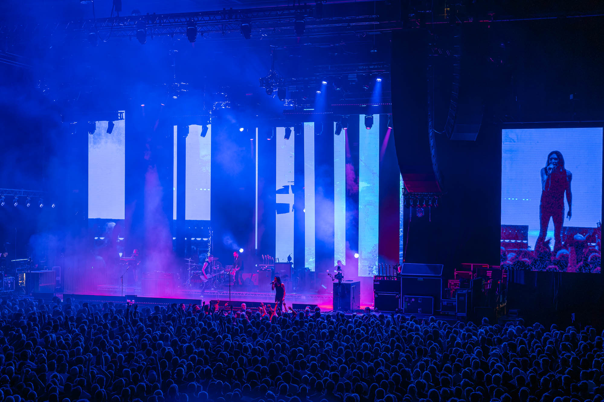 Brian Molko chantant au bord de la scène dans une lumière bleue et rose, écrans verticaux en fond, grand écran latéral montrant son image, public les bras levés.