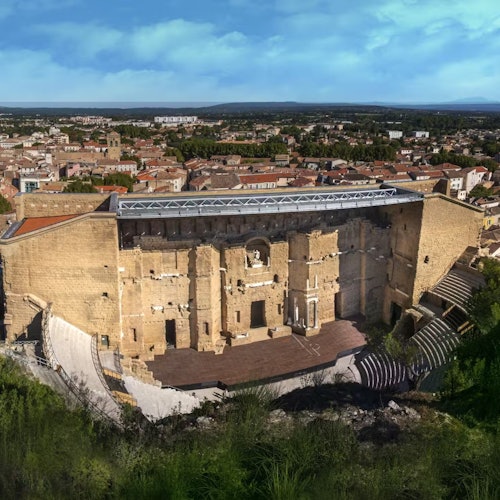 Đấu trường La Mã cổ đại với những bức tường đá lớn, hàng ghế xếp tầng và một sân khấu, đặt trong khung cảnh của một thị trấn và bầu trời xanh.