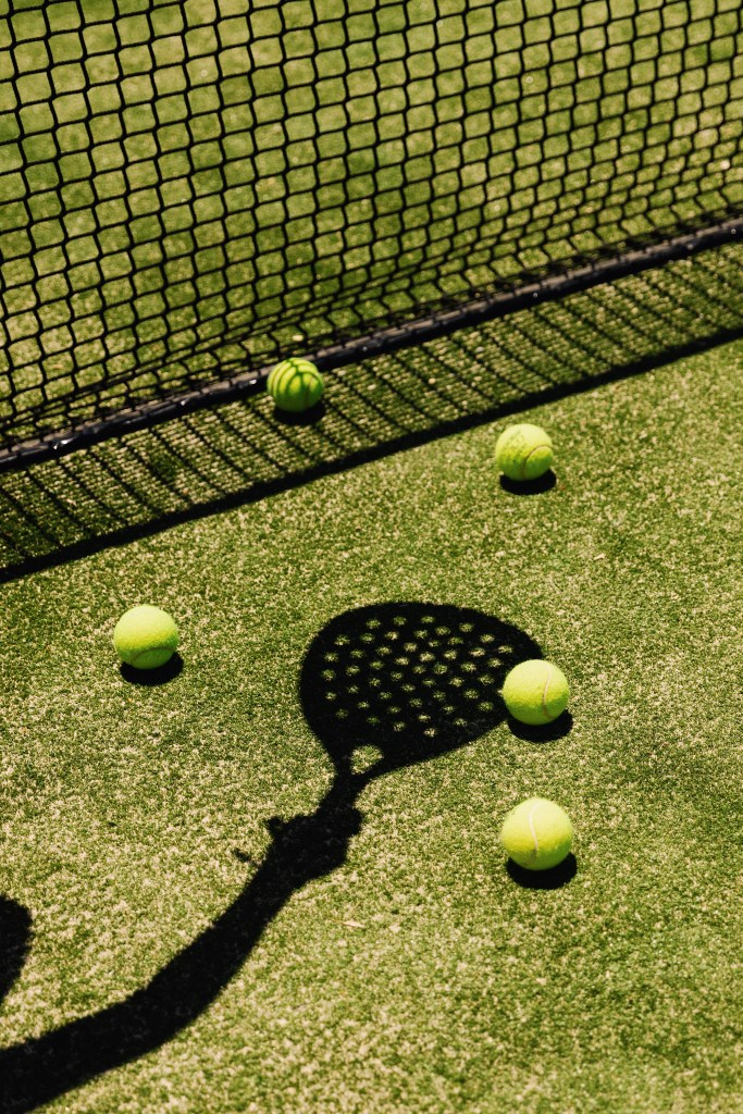 Shadow of a paddle racket on a green court with several yellow tennis balls scattered around.