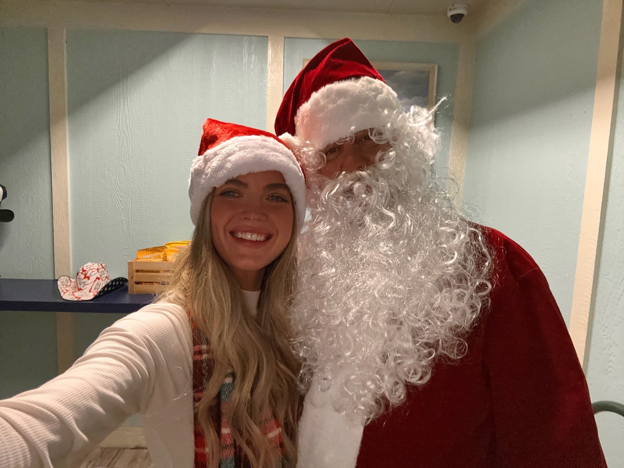 A person in a Santa hat smiles warmly next to someone dressed as Santa Claus, featuring a bright red suit and white beard, inside a festive room with light blue walls.