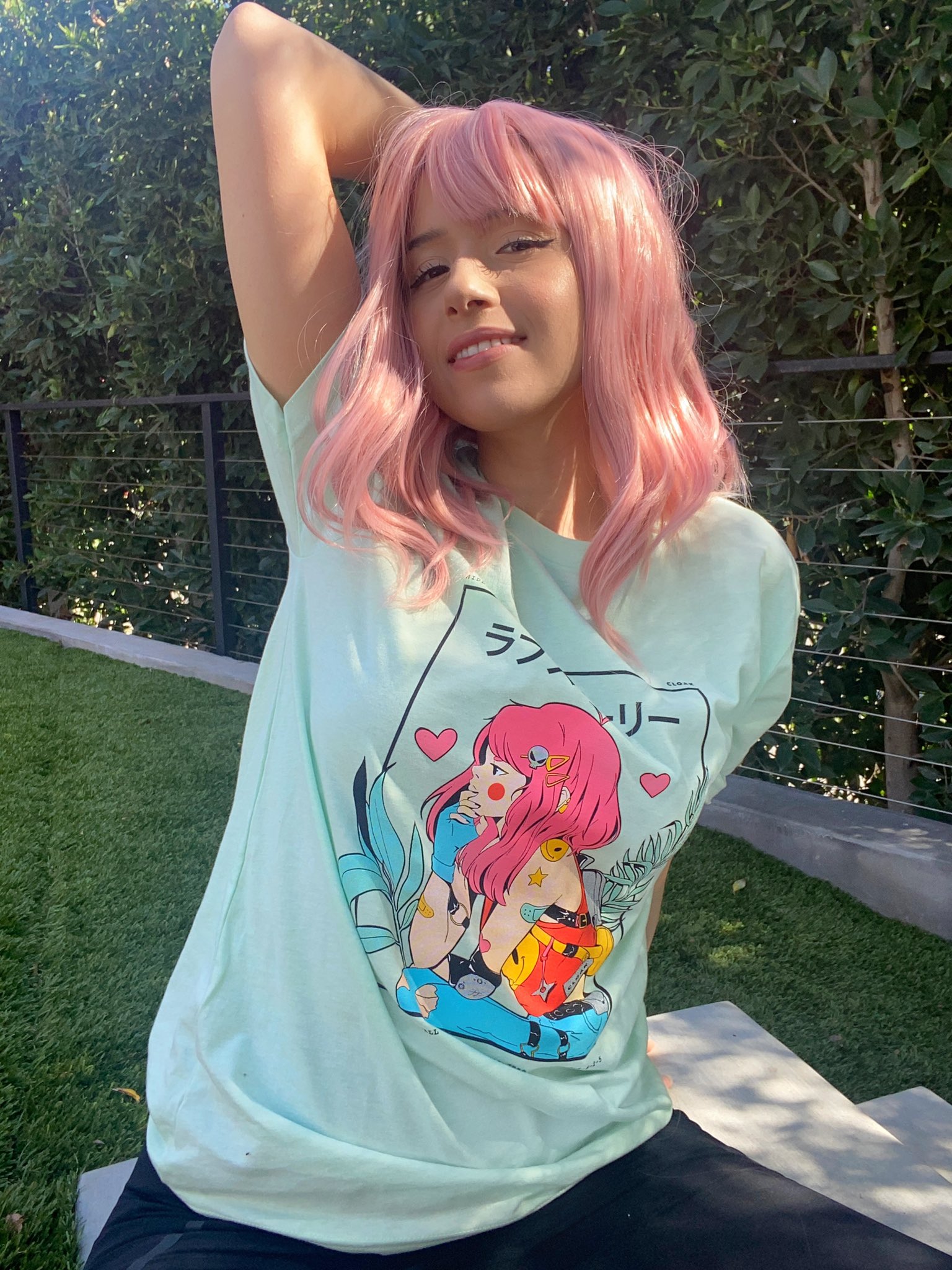 A person with pink hair poses playfully outdoors, wearing a light blue t-shirt with a colorful graphic design.