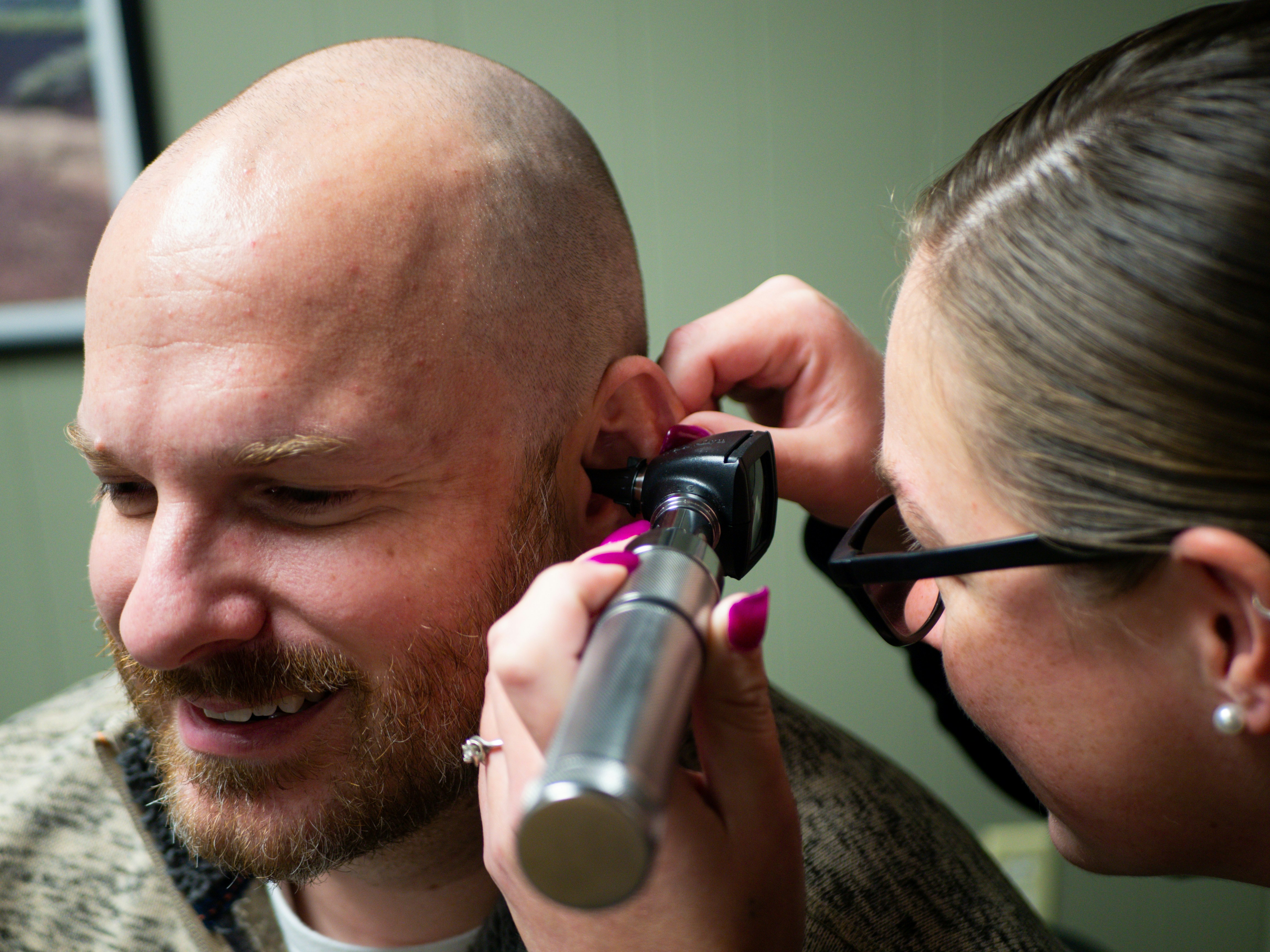 A person with a bald head smiles while another person examines their ear with a medical instrument.