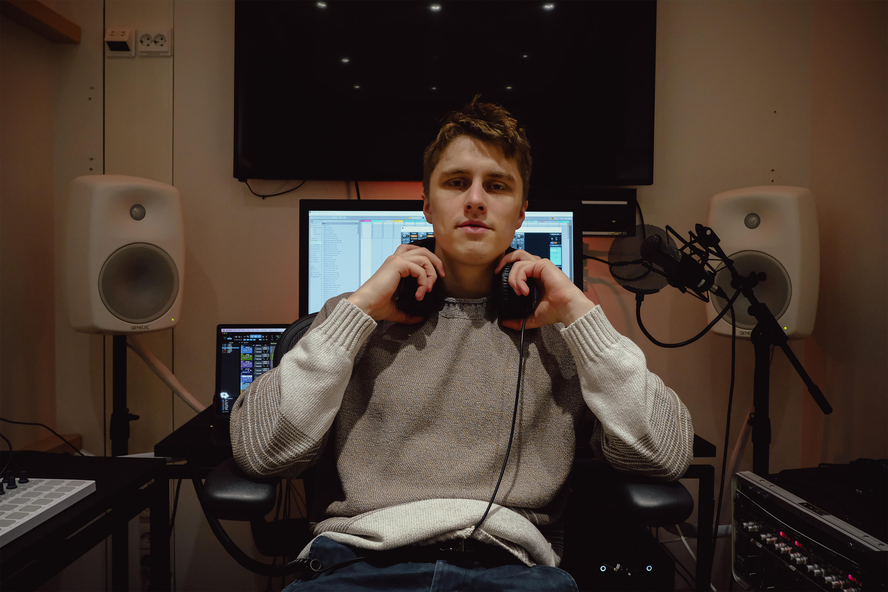 A person wearing headphones sits in a studio, focusing on a computer screen with audio equipment in the background.