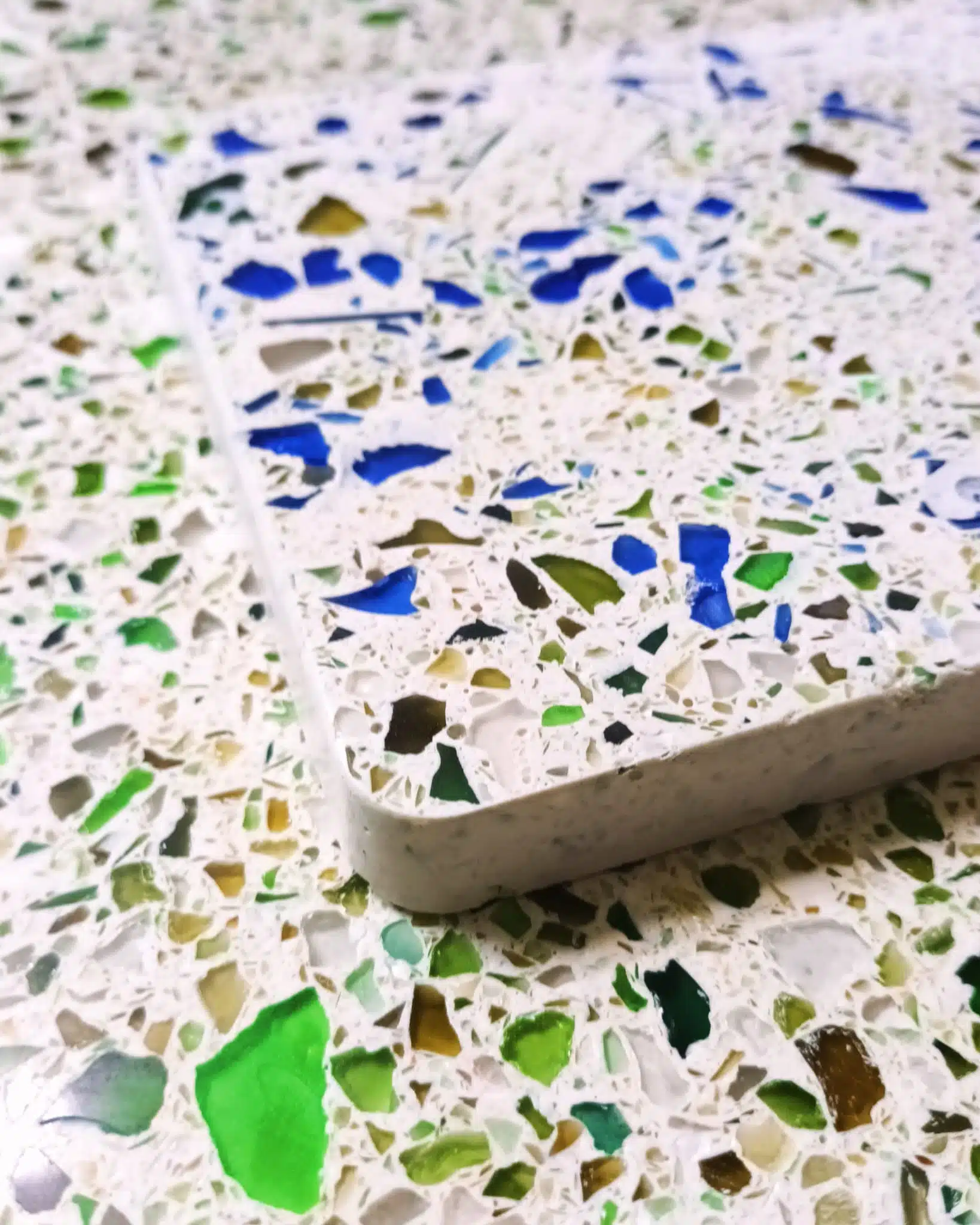 Gros plan d'une surface en terrazzo blanc incrustée d'éclats de verre recyclé aux teintes bleues, vertes et dorées, avec un carreau similaire posé dessus en angle