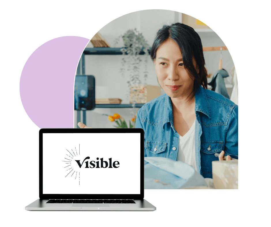 A woman in a casual denim shirt appears engaged in a video call, seated behind a laptop displaying the word "Visible" on its screen, set against a backdrop of home office decor including shelves and potted plants.