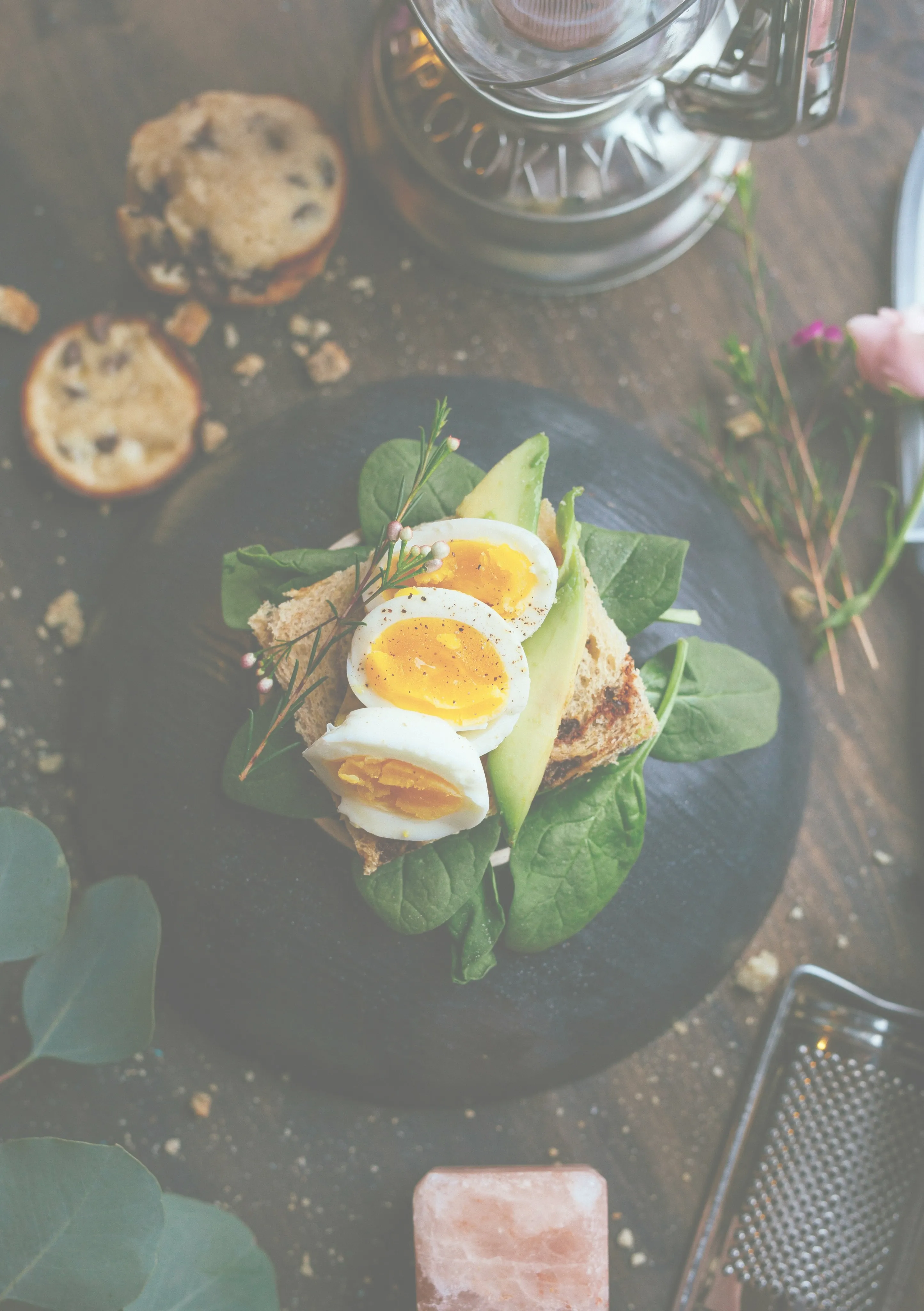 Open sandwich with boiled eggs, avocado slices, spinach, and herbs on dark bread, placed on a black plate. Rustic setting with cookies nearby.