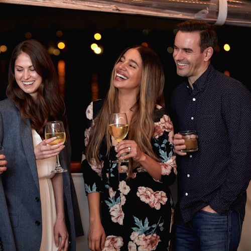 Three smiling people holding drinks, enjoying a night out. Background blurred with lights. One man, two women in casual attire.