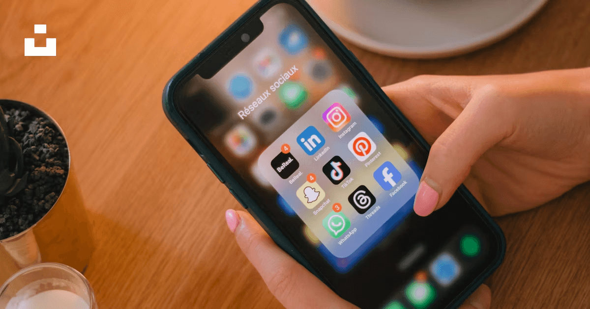 A close-up of someone holding a smartphone with a “Social Networks” folder open, showing apps like Instagram, LinkedIn, TikTok, Facebook, and WhatsApp.