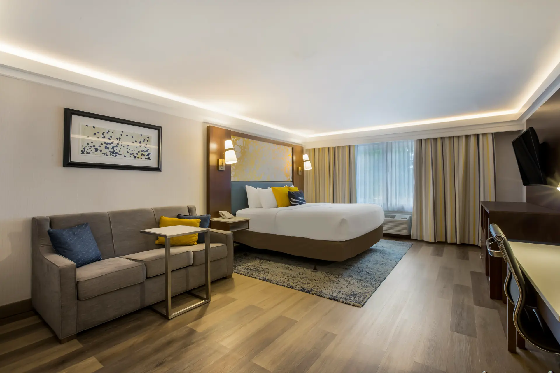 Modern hotel room with a large bed, desk, and kitchenette.