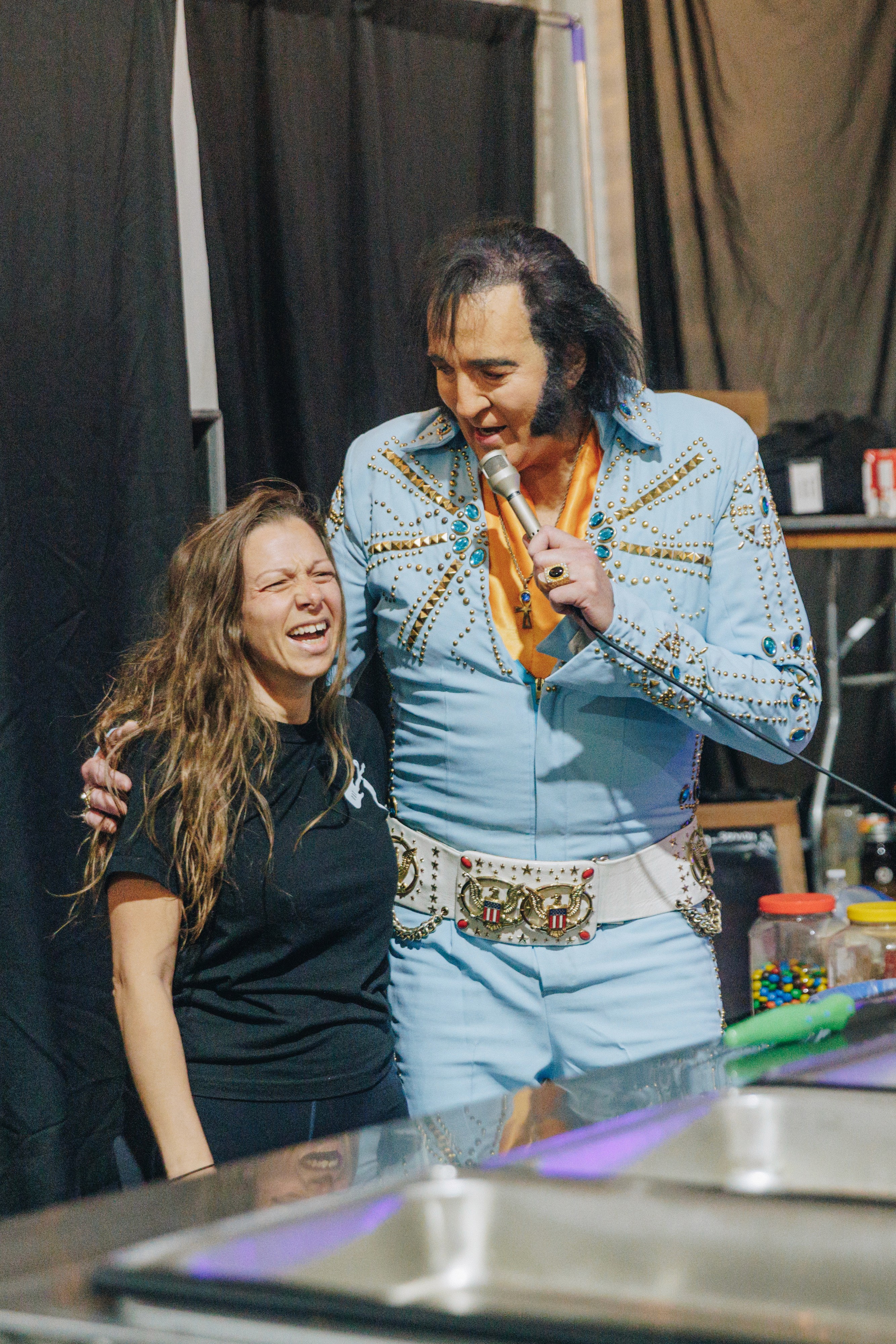 A smiling woman and a man in a blue outfit stand together, engaging in conversation at an event.