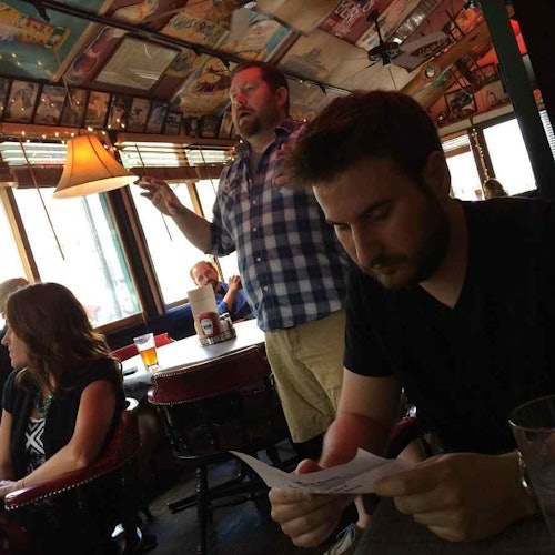 一名男子在桌子旁閱讀報紙，而另一名男子則在昏暗的餐廳中手勢表達，周圍還有幾位坐著的顧客。