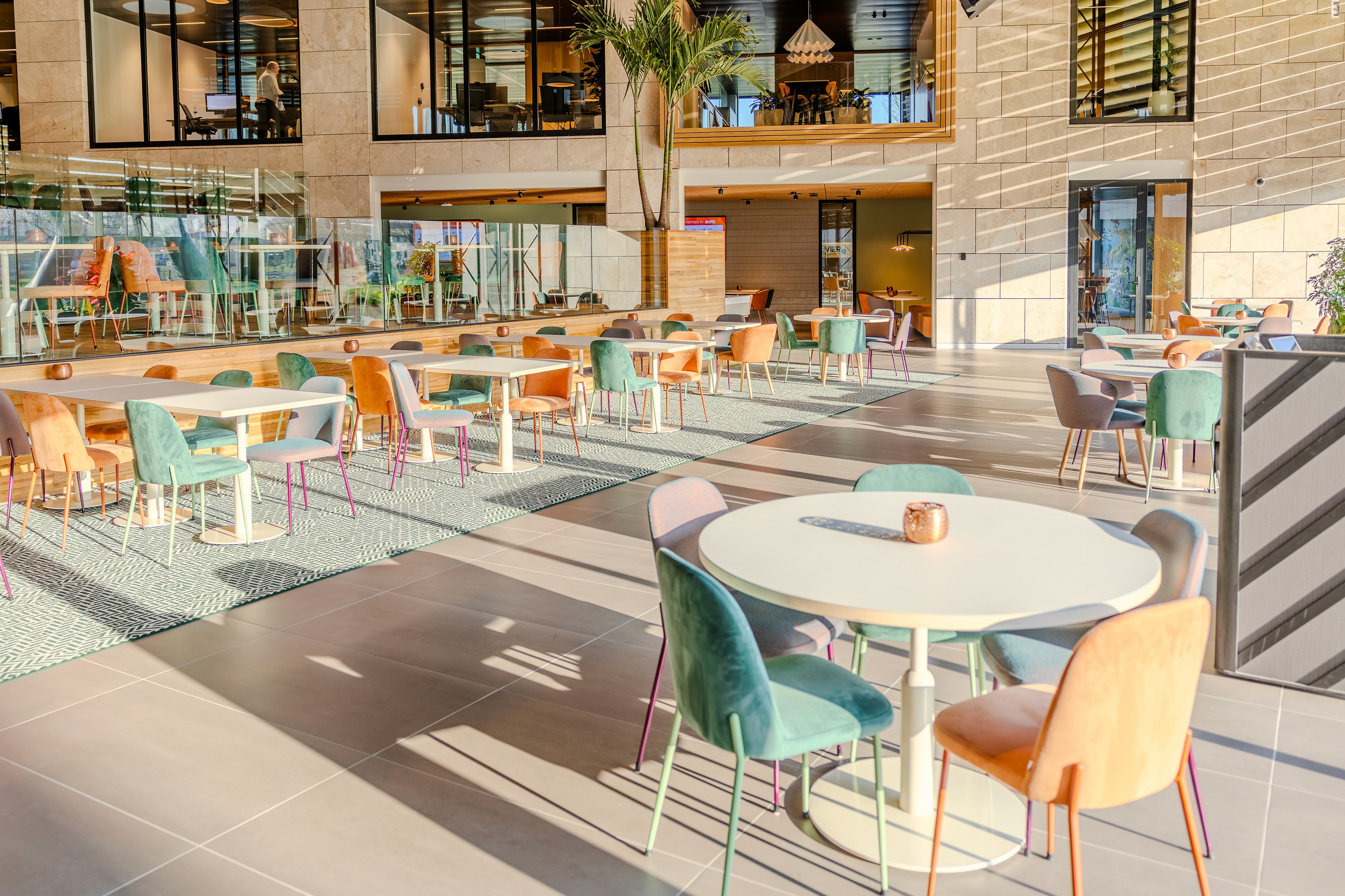 Ruime restaurant- en vergaderzone in AFAS DotNL met ronde witte tafels, groene en oranje stoelen op een geometrisch tapijt, omgeven door grote raampartijen